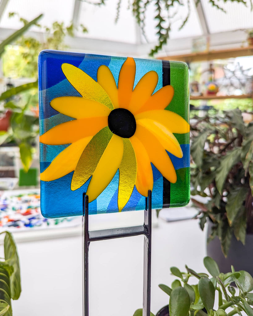 Fused glass miniature from the 'Abundance' series, featuring a bold yellow and orange sunflower with a black centre on a background of blue, green, and aqua, displayed on a black metal stand in a bright studio filled with plants