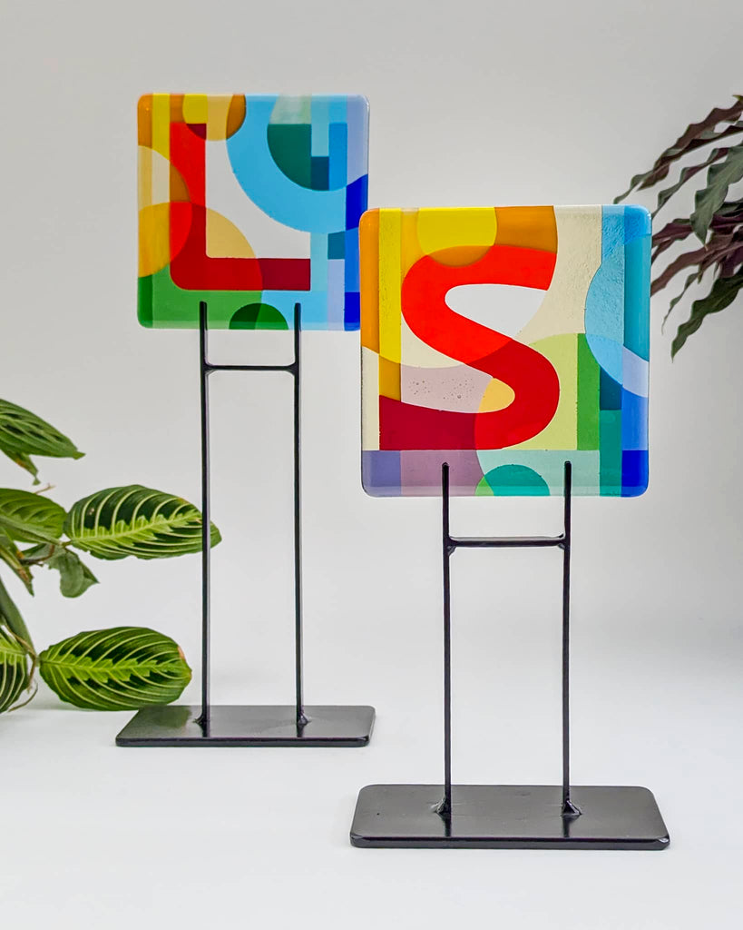 Pair of fused glass alphabet miniatures, featuring bold overlapping letters 'L' and 'S' in red, blue, yellow, and green, mounted on black metal stands