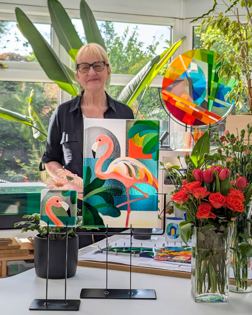 Artist Linda Rossiter in her glass studio, standing behind two fused glass artworks featuring flamingos and surrounded by tropical plants, colourful roundel art, and fresh cut flowers