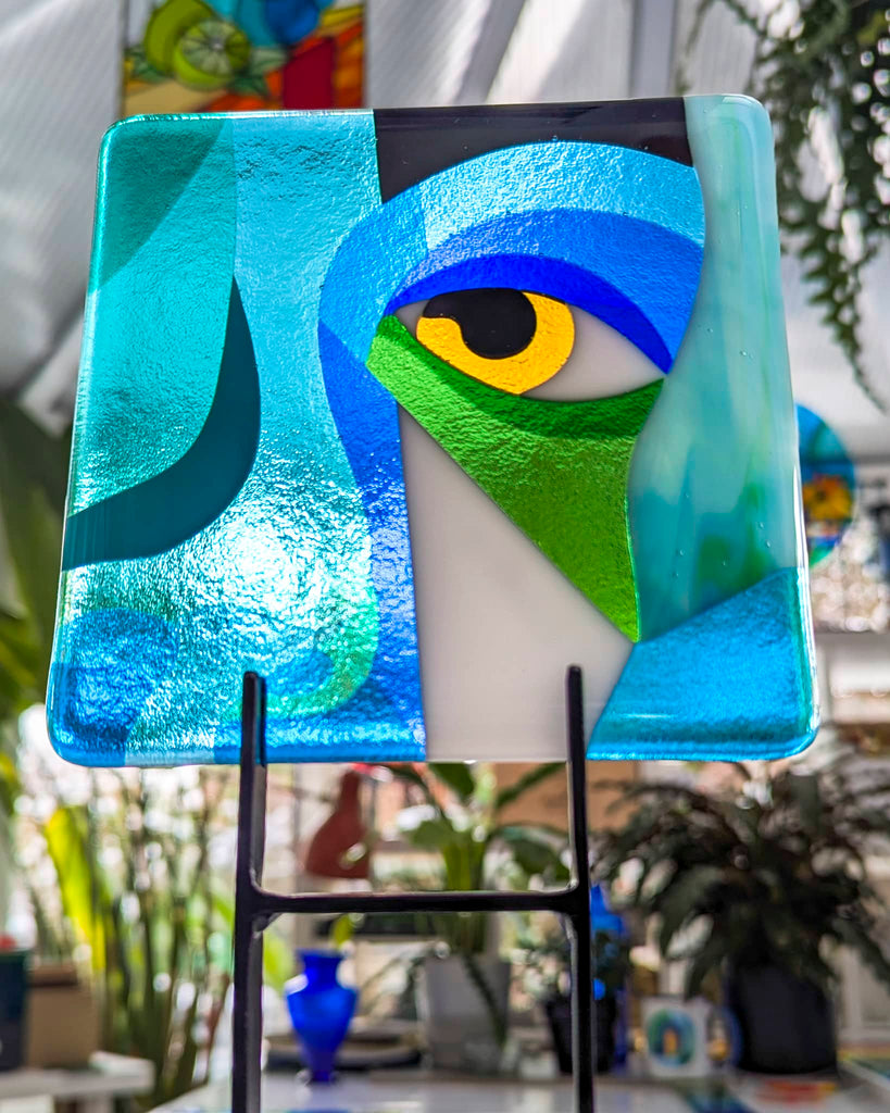 Fused glass artwork titled 'Bright Eyes', featuring a stylised yellow eye set within layered blue, green, and teal geometric forms, mounted on a black stand with a lush studio background