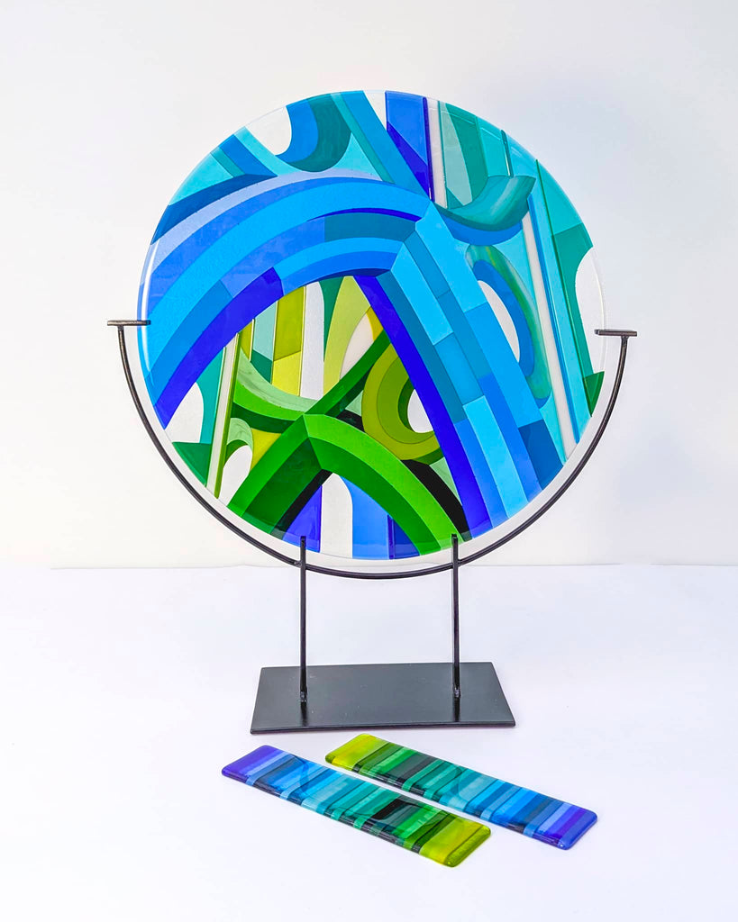 'Cobalt Cathedral' fused glass art - Round fused glass artwork titled 'Cobalt Cathedral', featuring geometric arcs and Hard Edge patterns in shades of blue and green, displayed on a black metal stand with glass test pieces in front