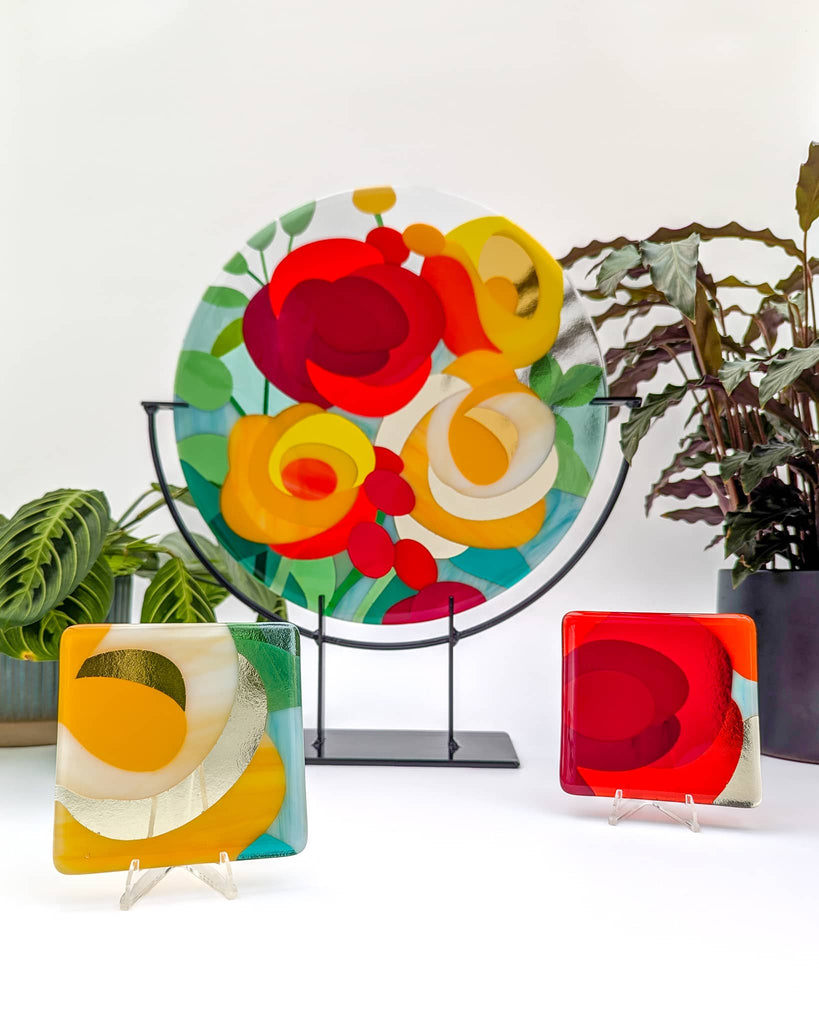 Fused glass roundel artwork with abstract floral design in bold red, yellow, orange, and white tones, displayed on a stand and accompanied by two matching miniature dishes, surrounded by lush green plants