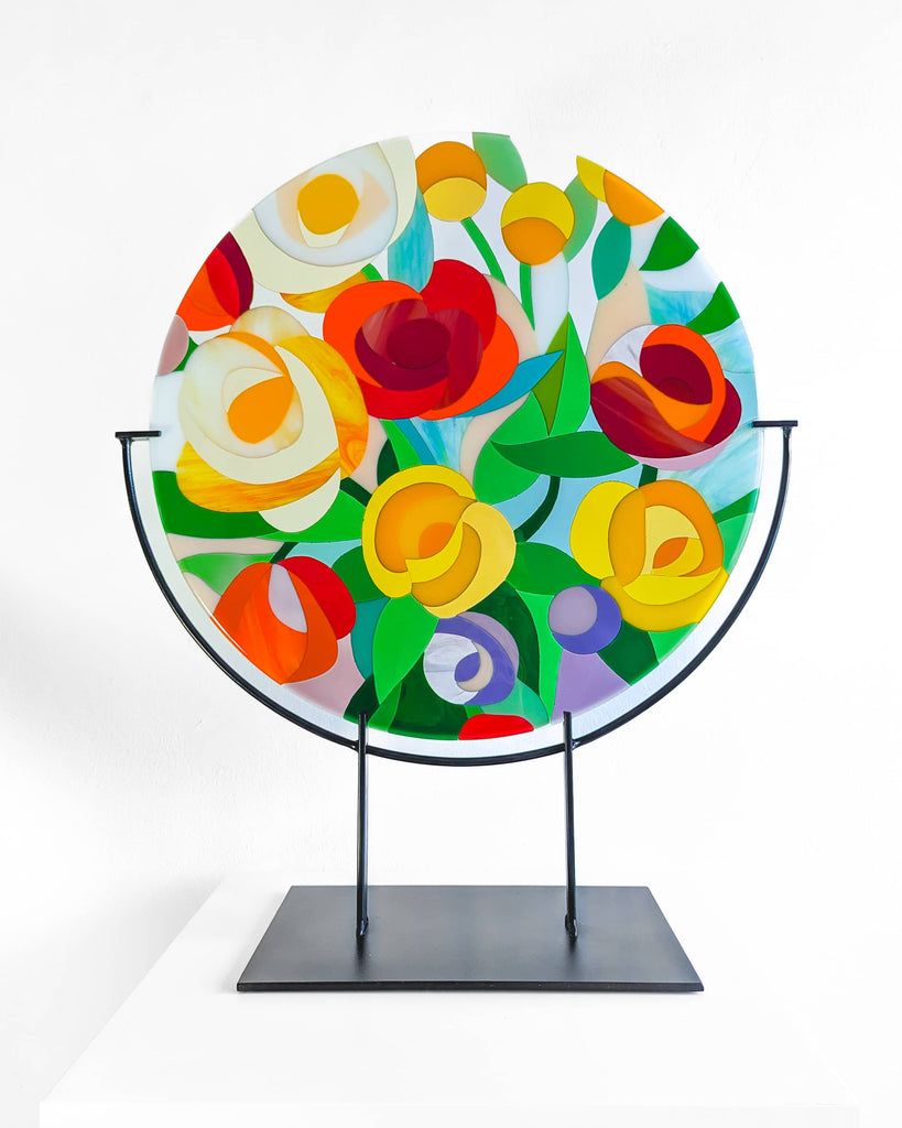 Floral-themed fused glass roundel featuring vibrant red, yellow, and orange petals, mounted on a black metal stand