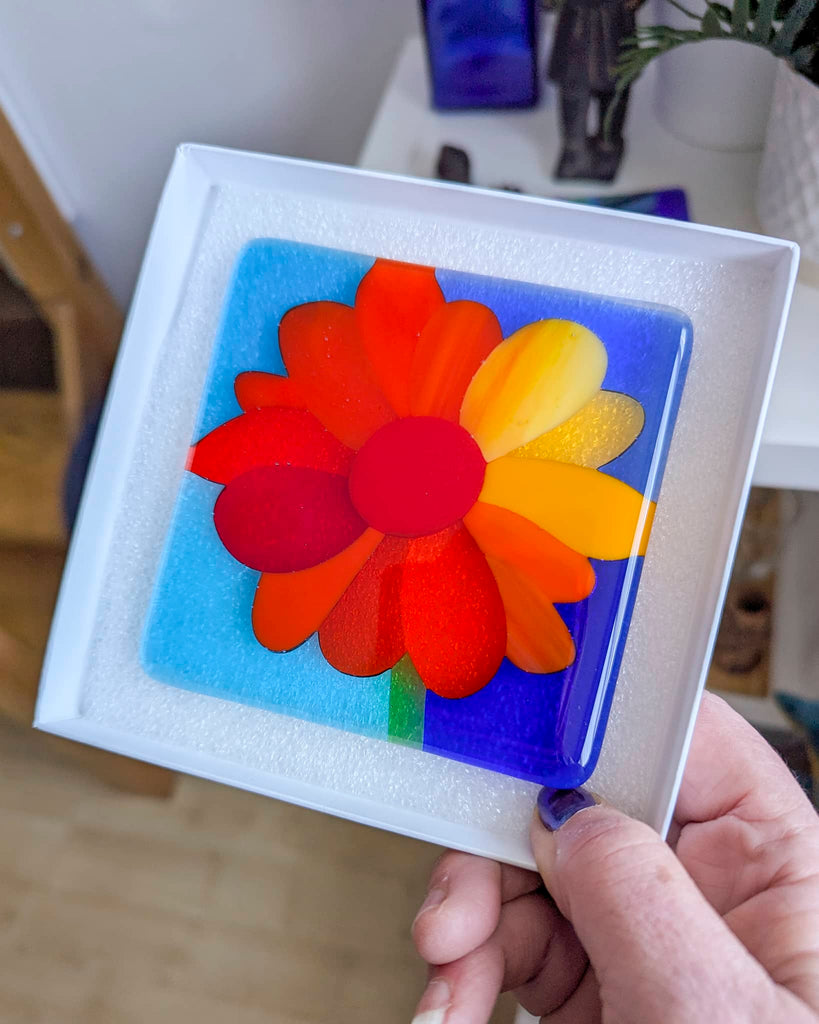 Square fused glass miniature featuring a bright red, orange, and yellow abstract flower on a blue and purple background, presented in a white gift box