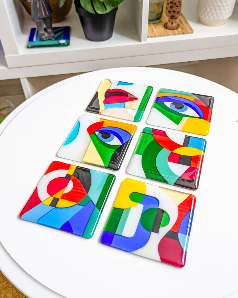 Set of six square fused glass miniatures from the 'Gaze' series, featuring abstract eyes, lips, and bold geometric shapes in vibrant primary colours, displayed on a white round table