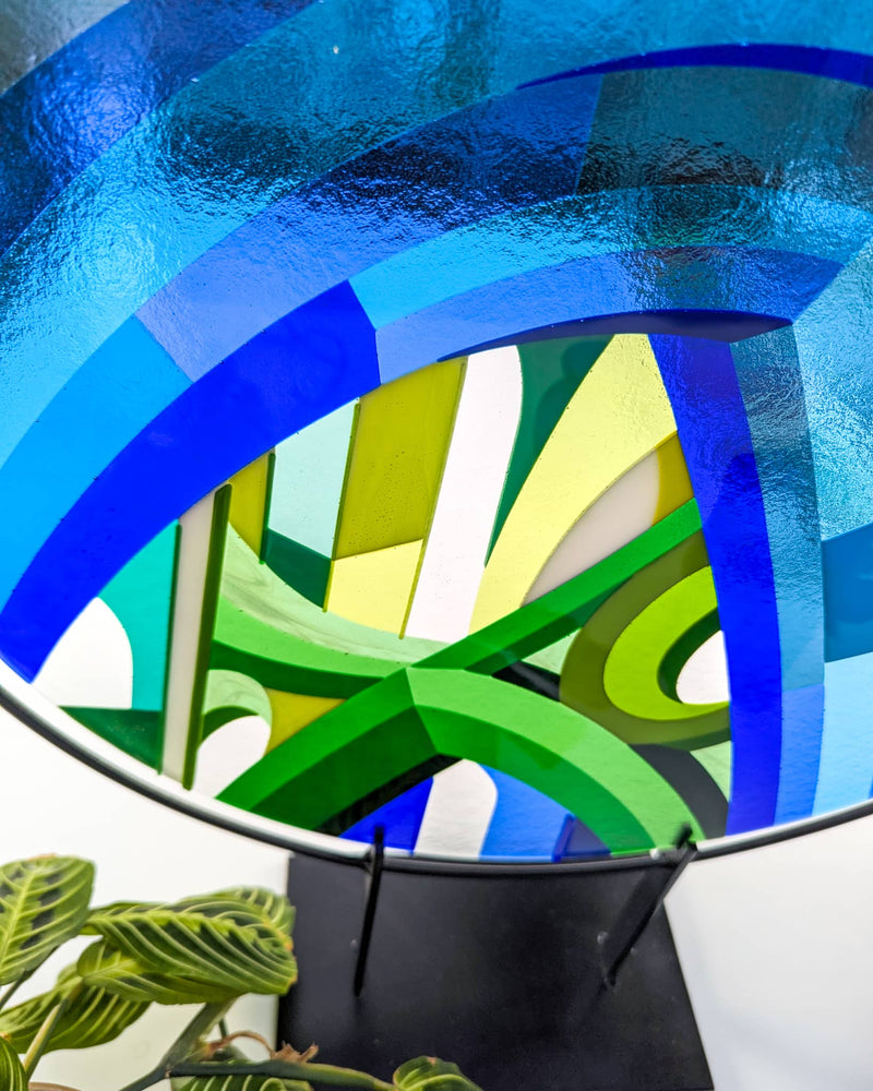Close-up of 'Cobalt Cathedral' fused glass art - Round fused glass artwork titled 'Cobalt Cathedral', featuring geometric arcs and Hard Edge patterns in shades of blue and green, displayed on a black metal stand