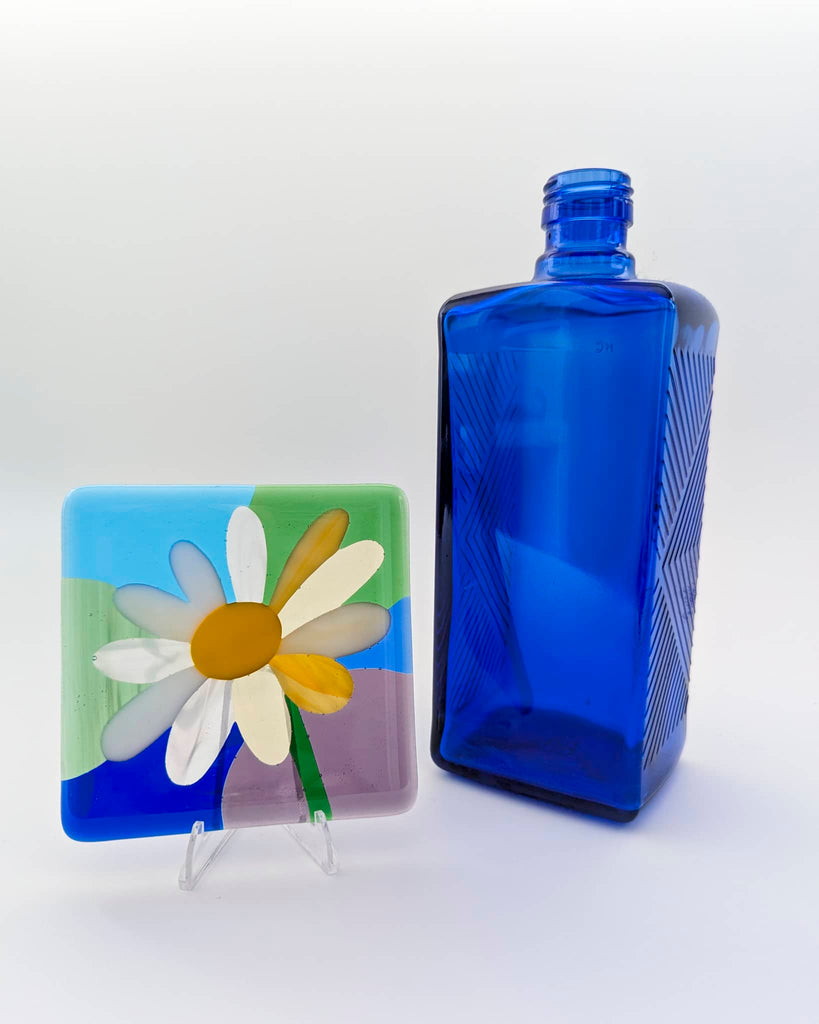 Fused glass miniature featuring a white and yellow daisy on a pastel background of blue, green, and lilac, displayed on a clear stand next to a cobalt blue glass bottle