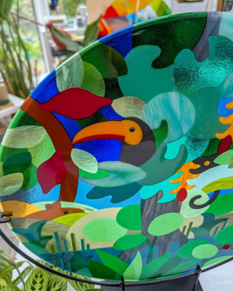 Hard Edge fused glass roundel with colourful toucan and tropical foliage, bespoke contemporary glass art on display stand