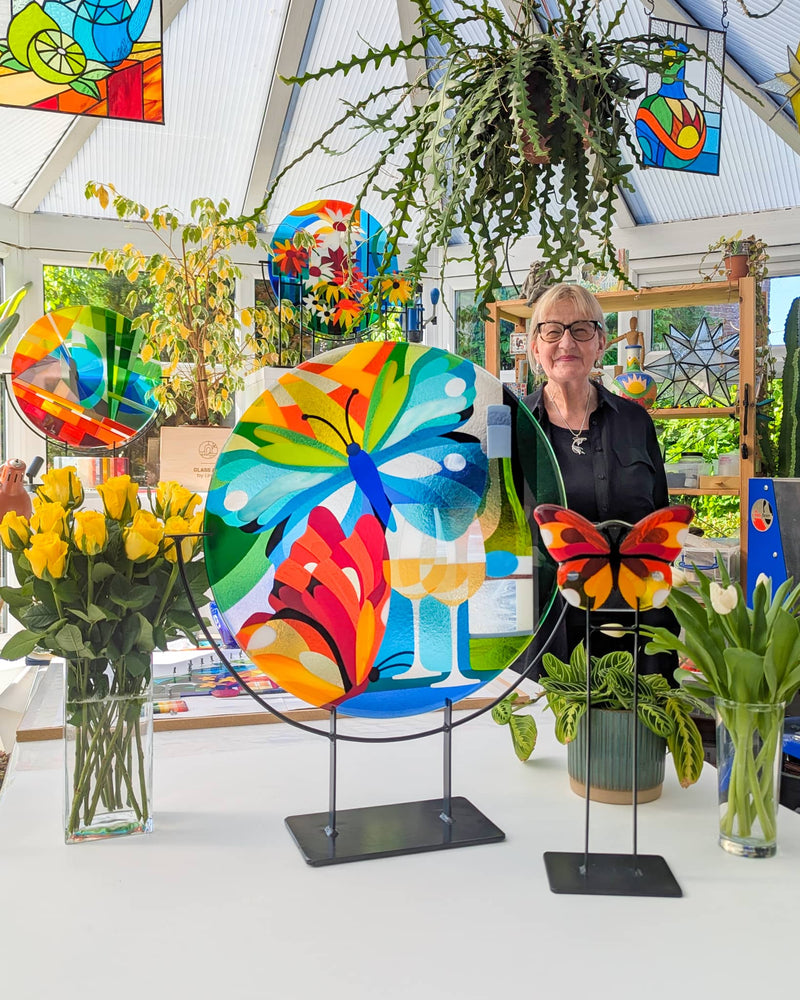 Vibrant fused glass artwork featuring butterflies, wine glasses, and colourful foliage on stands, displayed in a sunlit studio - a personal commission from Glass Art by Linda