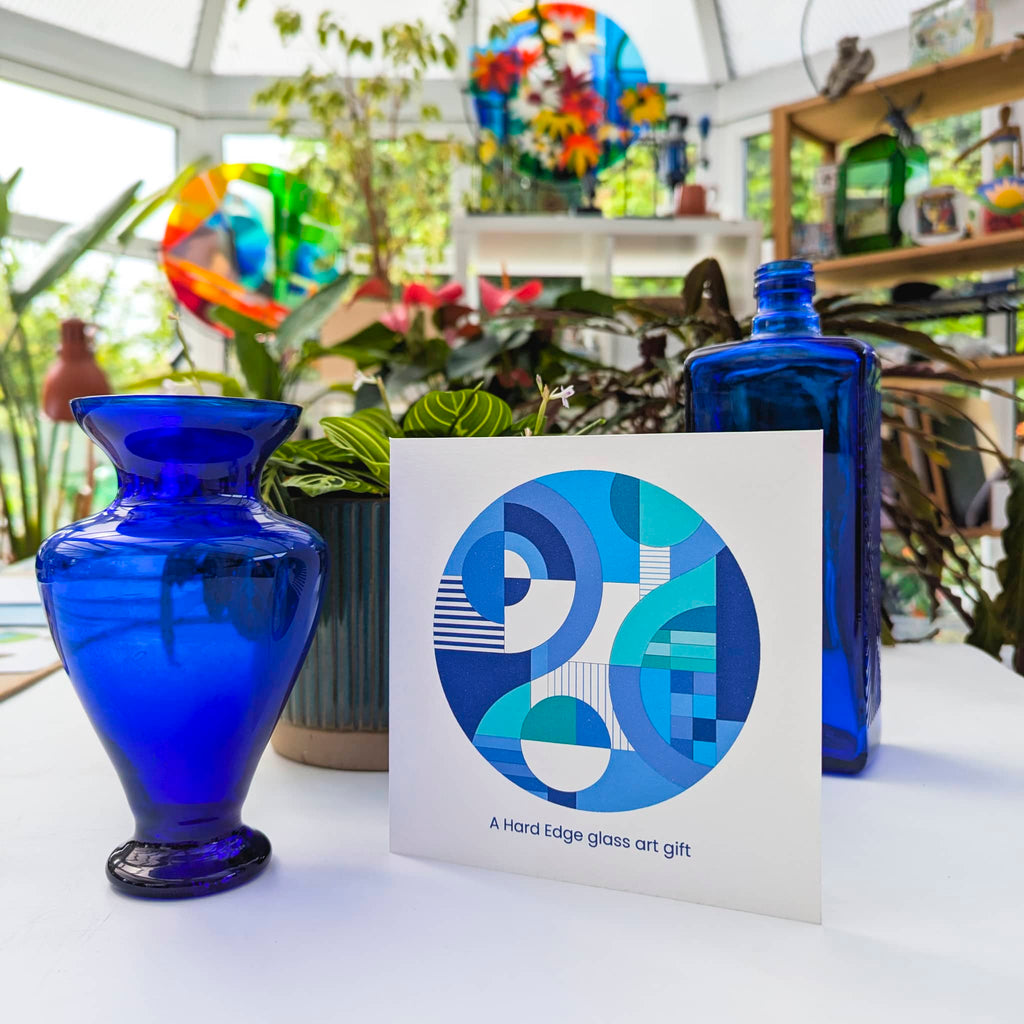 Gift card for a Hard Edge glass art commission, featuring a blue geometric design, displayed between cobalt blue glass vases in a bright, plant-filled studio.