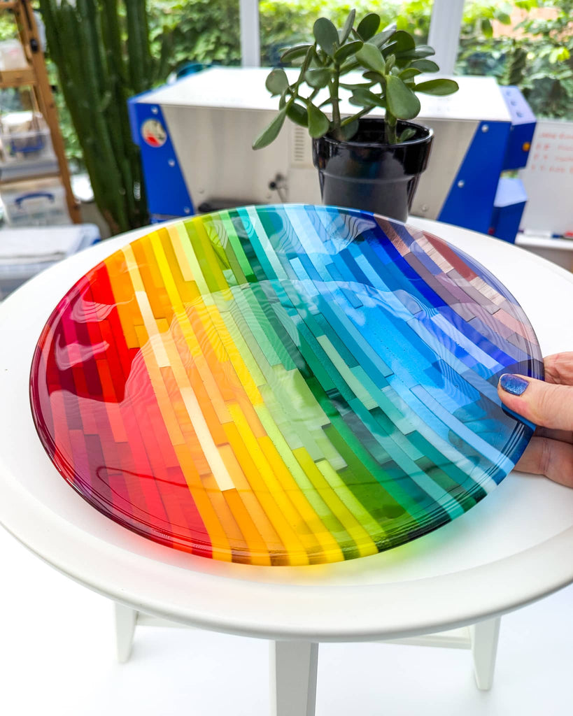 Fused glass art dish titled 'Rainbowl', featuring vibrant rainbow stripes in a linear Hard Edge pattern, held over a white table in a bright studio setting