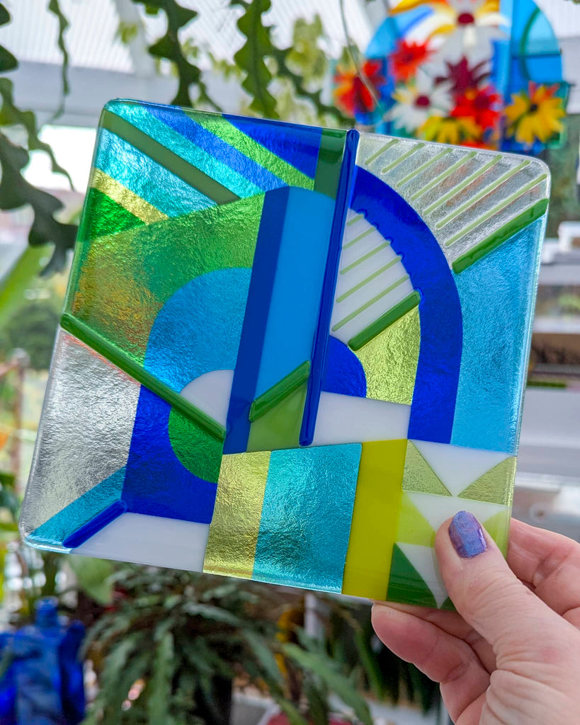 Square fused glass panel titled 'Spring Awakens', featuring vibrant geometric patterns in greens, blues, and yellows with layered detailing