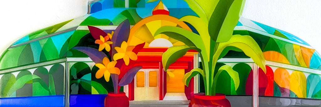 Detail of a vibrant fused glass artwork depicting a stylised glasshouse with red double doors, colourful foliage, and two large potted plants in the foreground. Layers of green, blue, yellow and orange glass form the arched structure