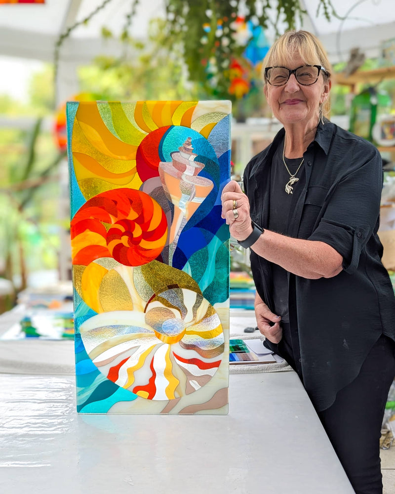 Linda Rossiter presenting completed Hard Edge fused glass panel featuring nautilus shells and abstract figure, bespoke contemporary glass art commission