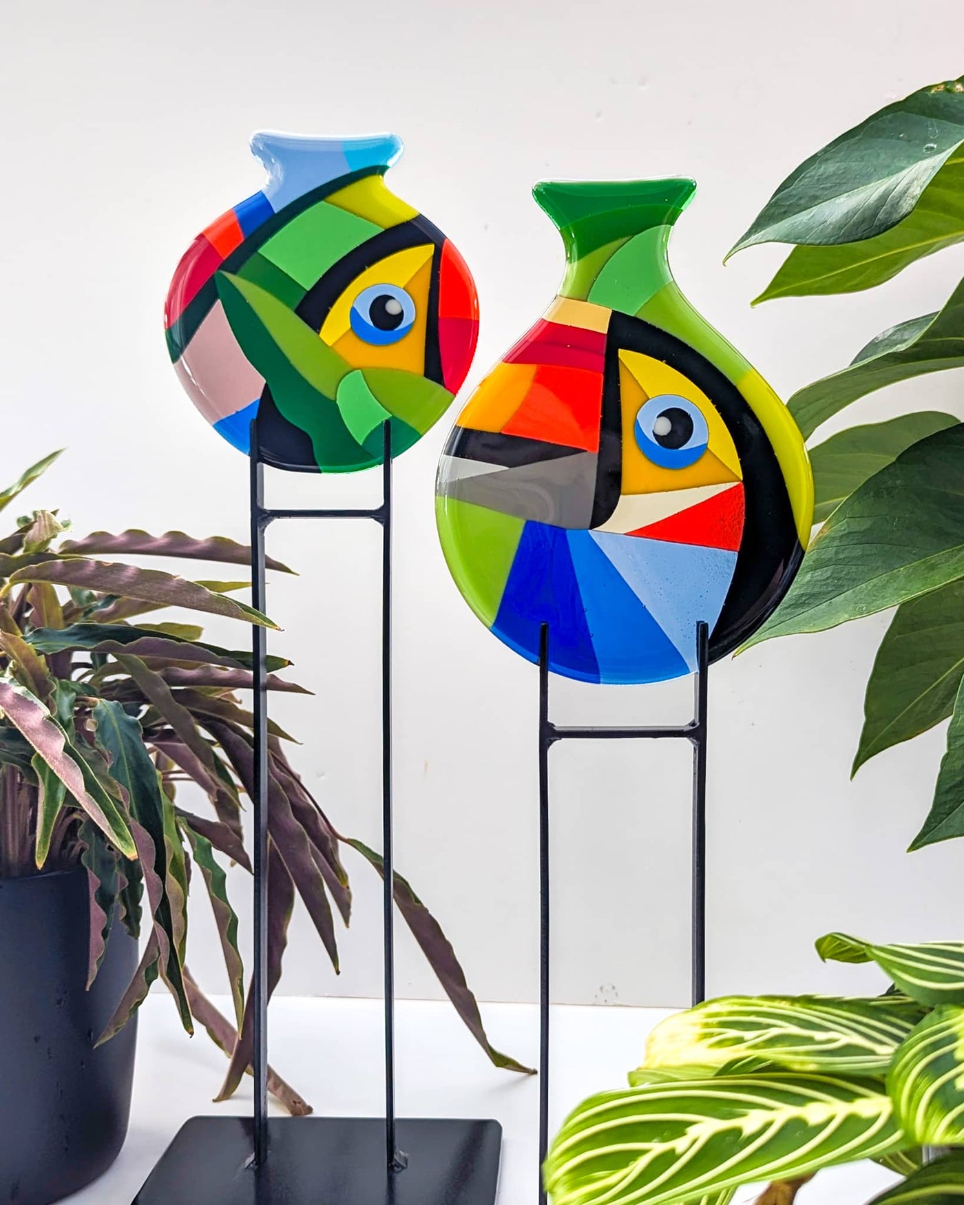 Close-up of the two 'Toucans' contemporary Hard Edge glass miniatures on their steel stands, showing the bold geometric forms and contrasting colour palettes that complement the larger roundel