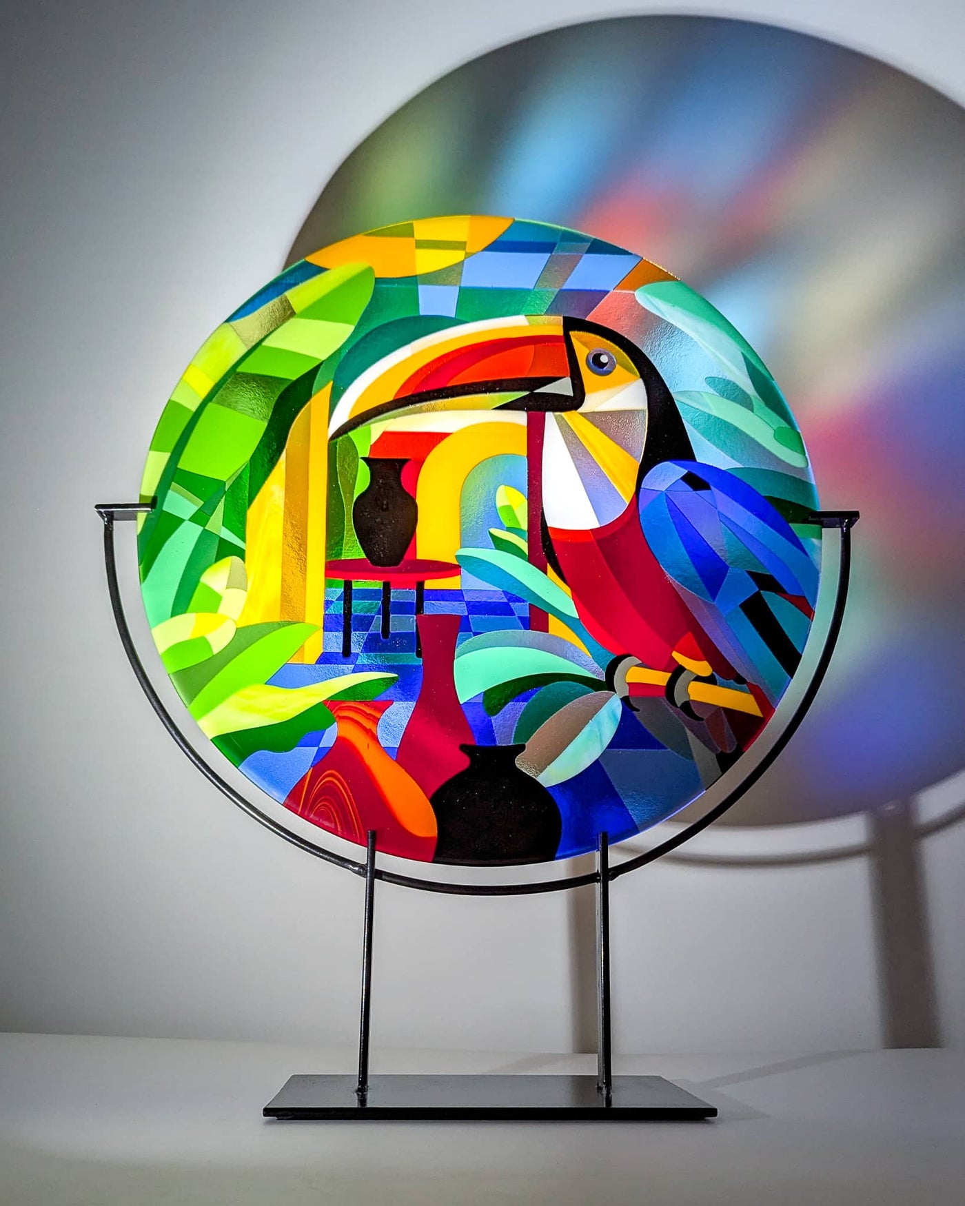 'Toucans' contemporary Hard Edge glass roundel by Linda Rossiter displayed on its steel stand, depicting a colourful toucan surrounded by tropical foliage and vibrant geometric forms