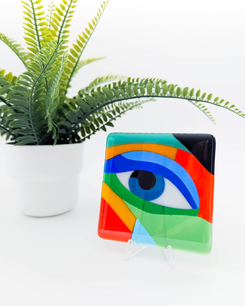 Fused glass miniature artwork titled 'Yearning', featuring a stylised blue eye within abstract geometric shapes in red, green, black, orange, and blue, displayed on a clear stand