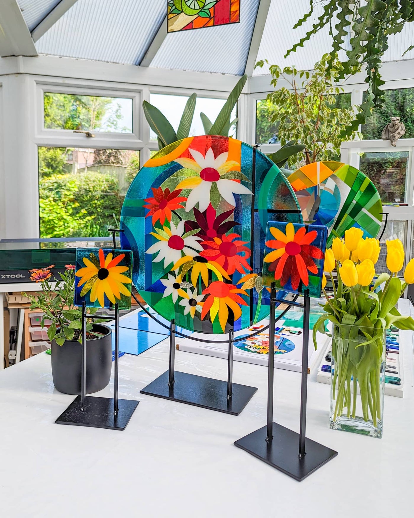'Abundance' contemporary Hard Edge glass roundel with two matching floral miniatures on steel stands, featuring a vibrant arrangement of colourful flowers