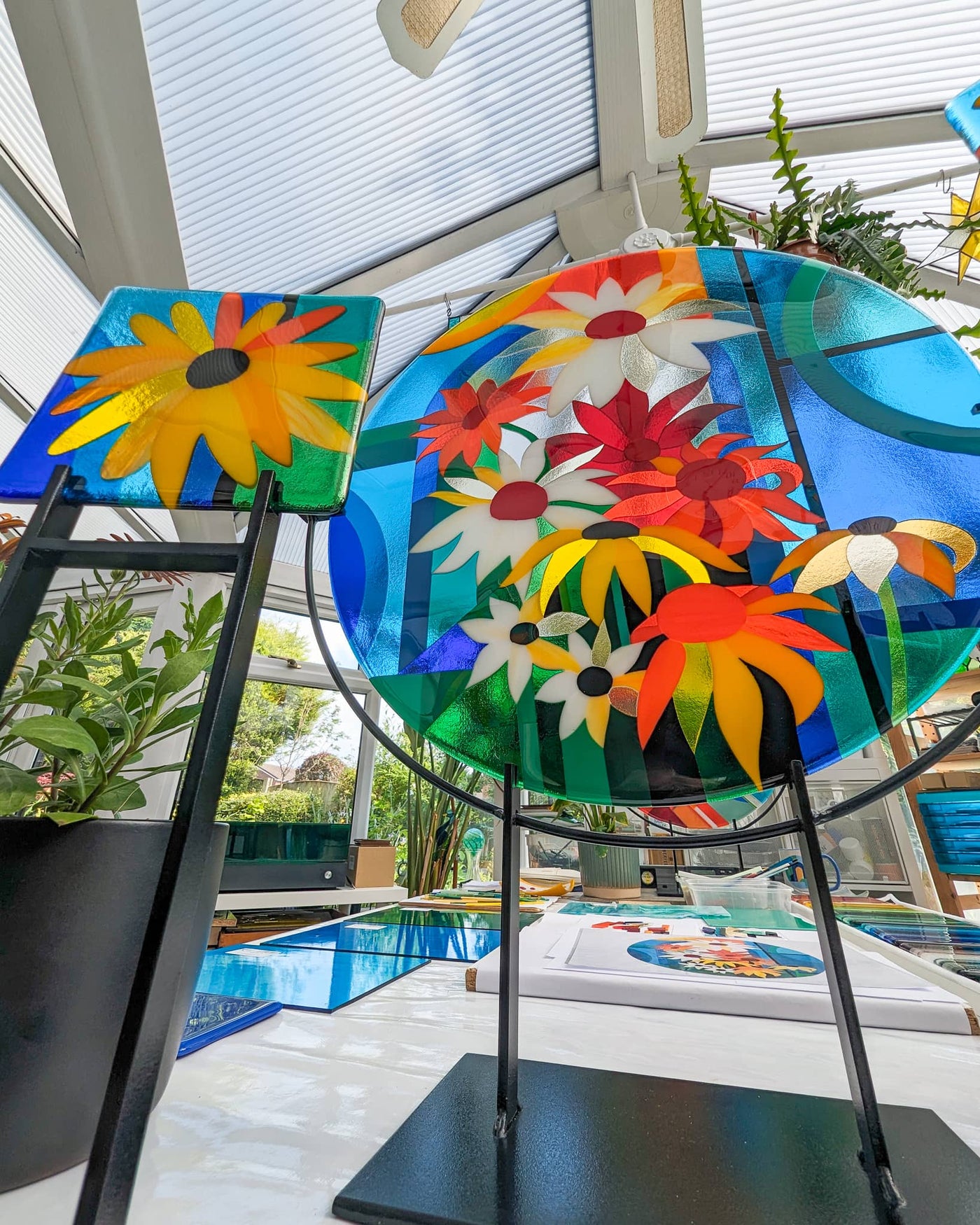 'Abundance' contemporary Hard Edge glass roundel displayed with one of the matching floral miniatures on steel stands in Linda Rossiter's studio