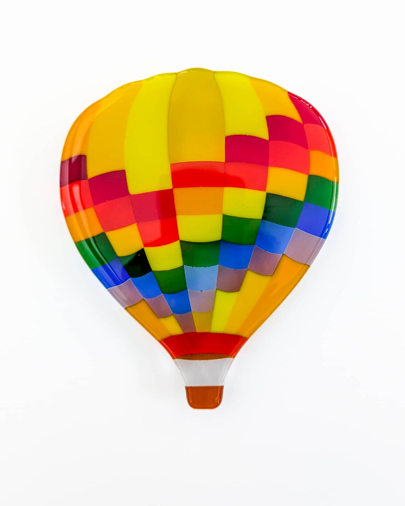 Fused glass artwork in the shape of a hot air balloon, featuring a vibrant patchwork of rainbow colours including red, yellow, green, blue, and purple on a white background