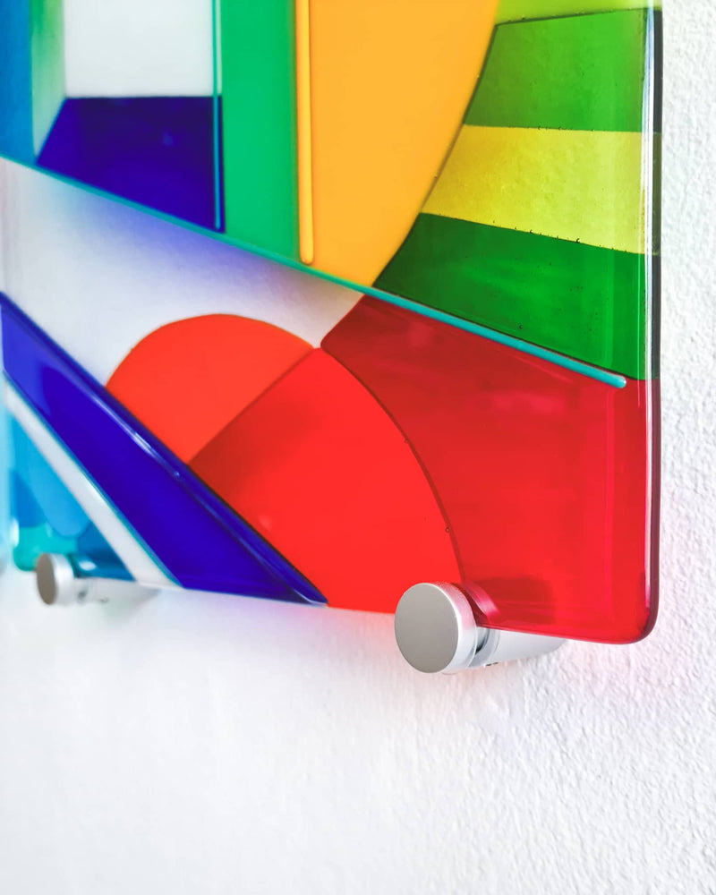 Close-up of the Biarritz glass wall art showing red, green, blue and yellow shapes held away from the wall by a metal fixing