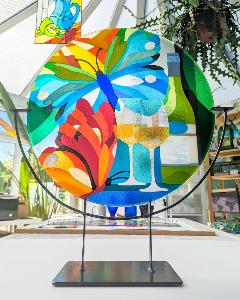 Hard Edge fused glass roundel artwork with colourful butterflies and wine glasses on a custom stand, contemporary glass art by Linda Rossiter