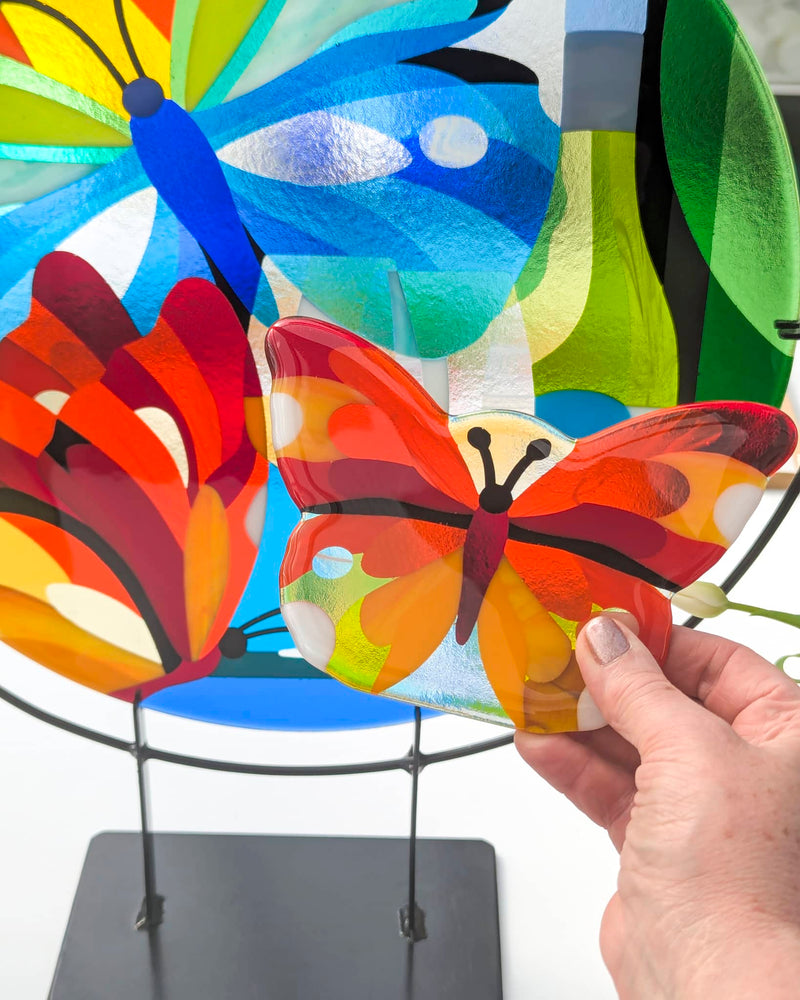 Close-up of vibrant fused glass artwork featuring butterflies, wine glasses, and colourful foliage on stands - a personal commission from Glass Art by Linda