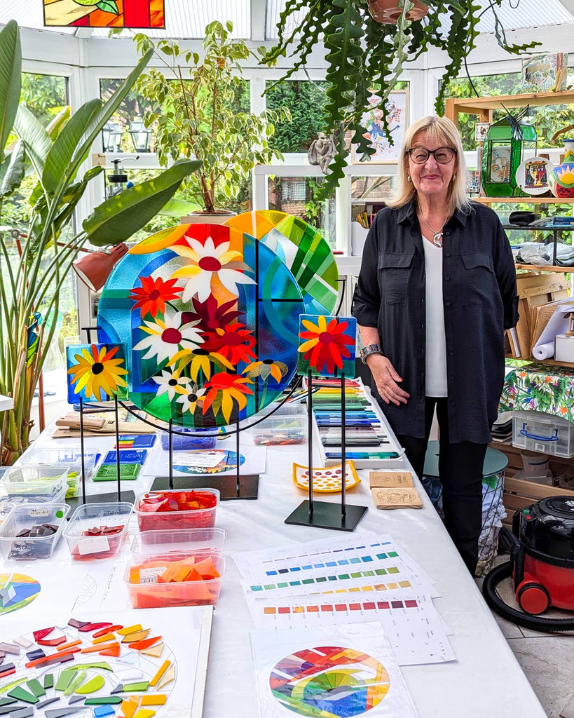 Glass artist Linda Rossiter standing in her bright studio surrounded by colourful fused glass artworks, design samples, and trays of coloured glass – ready to guide clients through the commissioning process