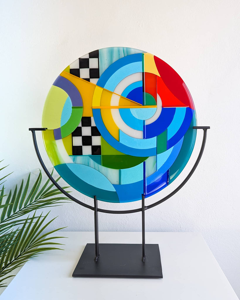 Fused glass roundel artwork titled 'Chequer', featuring bold Hard Edge geometric patterns in red, blue, yellow, green, and black-and-white chequerboard elements, displayed on a black metal stand