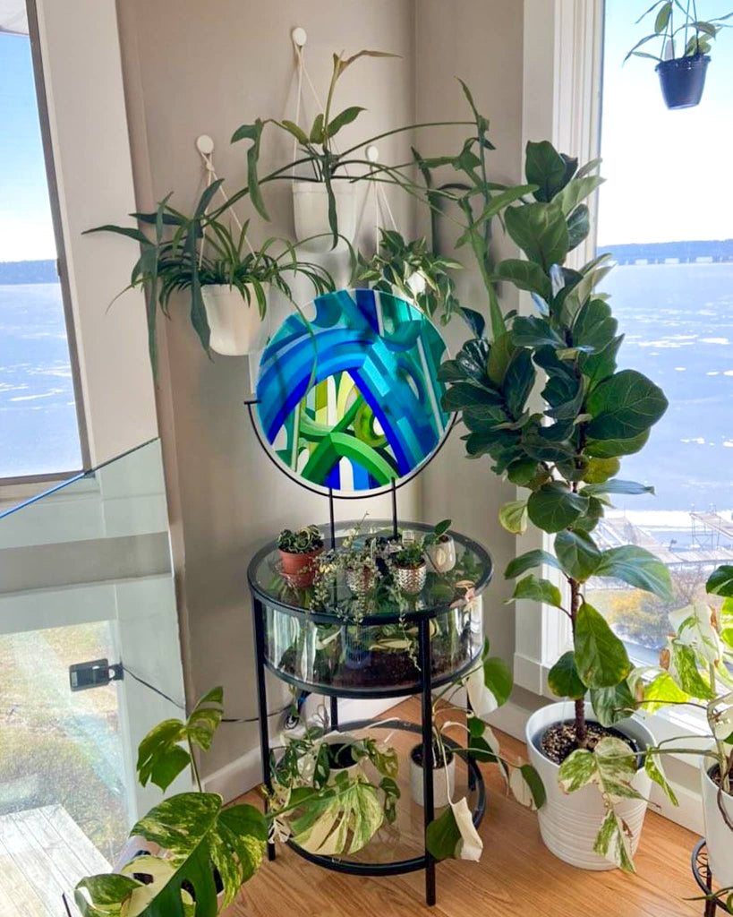 Fused glass artwork titled ‘Cobalt Cathedral’ displayed in a client’s home, set on a glass table surrounded by lush indoor plants with a view of the sea beyond – a commissioned piece from Glass Art by Linda