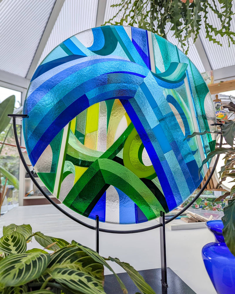 'Cobalt Cathedral' fused glass art - Round fused glass artwork titled 'Cobalt Cathedral', featuring geometric arcs and Hard Edge patterns in shades of blue and green, displayed on a black metal stand