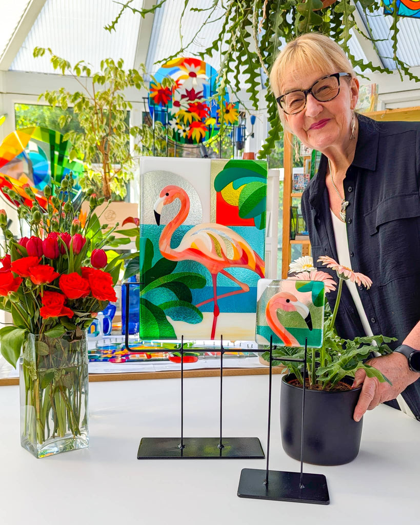 'Glass artist Linda Rossiter standing beside two flamingo-themed fused glass artworks on stands, surrounded by flowers in a bright, plant-filled studio'