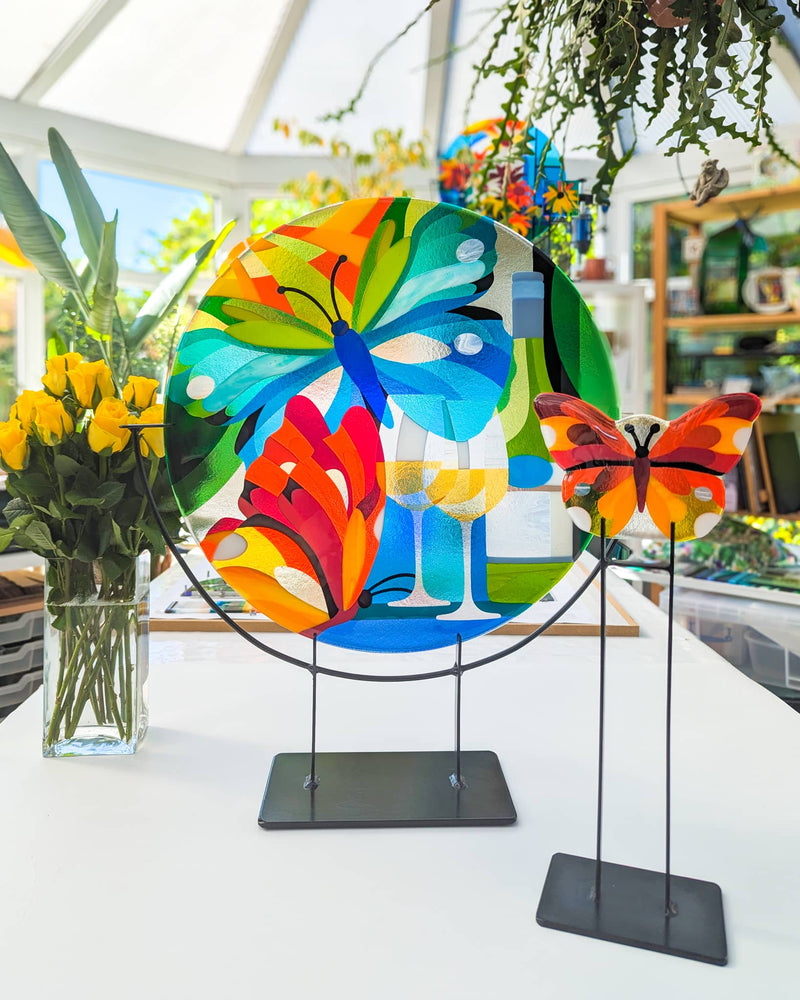 Vibrant fused glass artwork featuring butterflies, wine glasses, and colourful foliage on stands, displayed in a sunlit studio – a personal commission from Glass Art by Linda