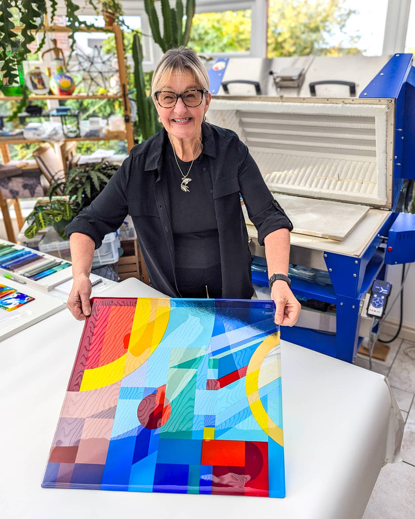 Glass artist Linda Rossiter smiling in her studio while holding a newly completed Hard Edge fused glass panel in vibrant colours, with the kiln open behind her – marking the final reveal of a commissioned artwork