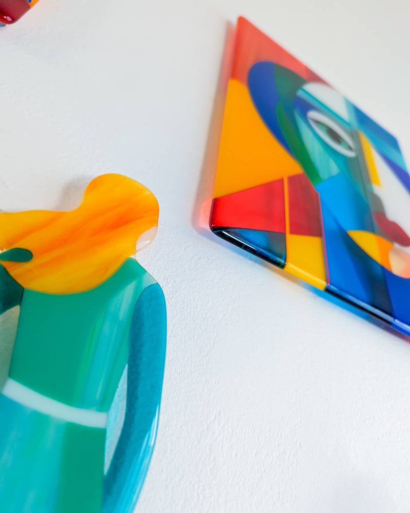 Angled view of a turquoise glass figure and the abstract portrait panel from Gallery mounted away from a white wall