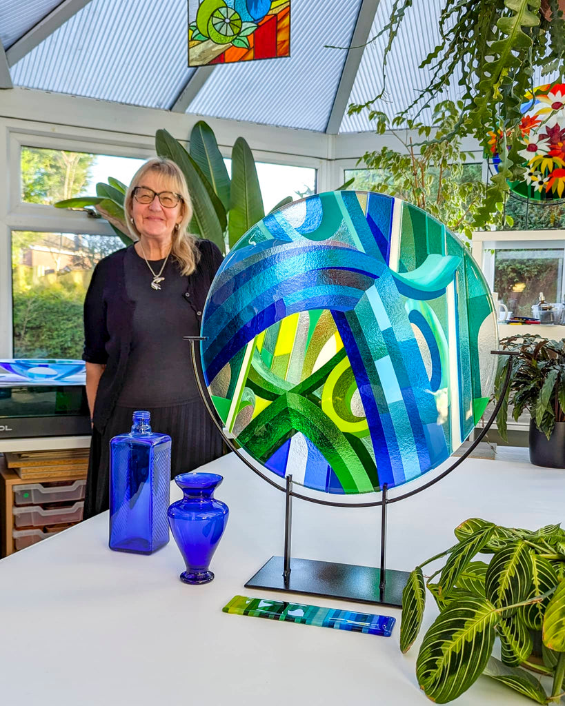 Glass artist Linda Rossiter standing beside a large round fused glass artwork in cool green and blue tones, displayed with cobalt blue vases in a bright studio setting