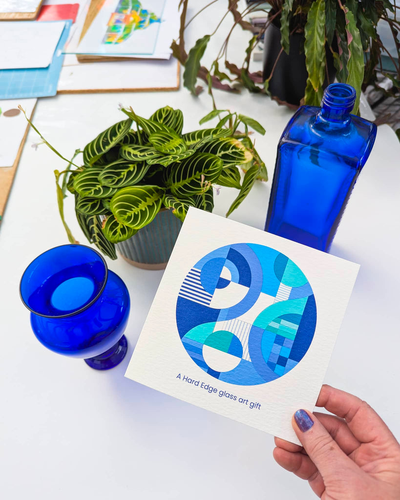 Hand holding a printed card featuring a blue-toned Hard Edge design, with vibrant cobalt glassware and a leafy plant on the table – highlighting accessible miniatures from Glass Art by Linda