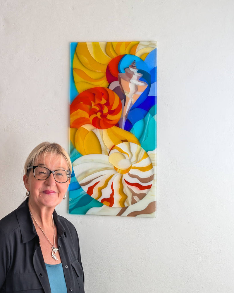 Linda Rossiter standing beside The Sea, a tall colourful fused glass wall panel featuring shells and conches