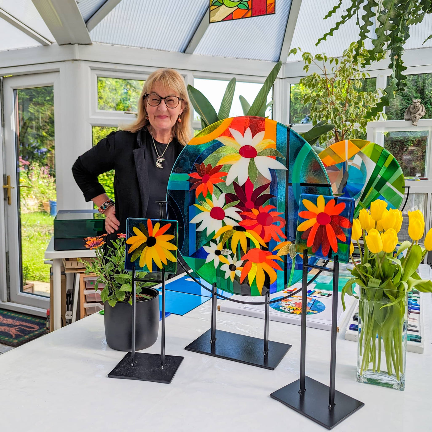 Linda Rossiter with the completed 'Abundance' contemporary Hard Edge glass roundel and two matching floral miniatures displayed on their steel stands in her studio
