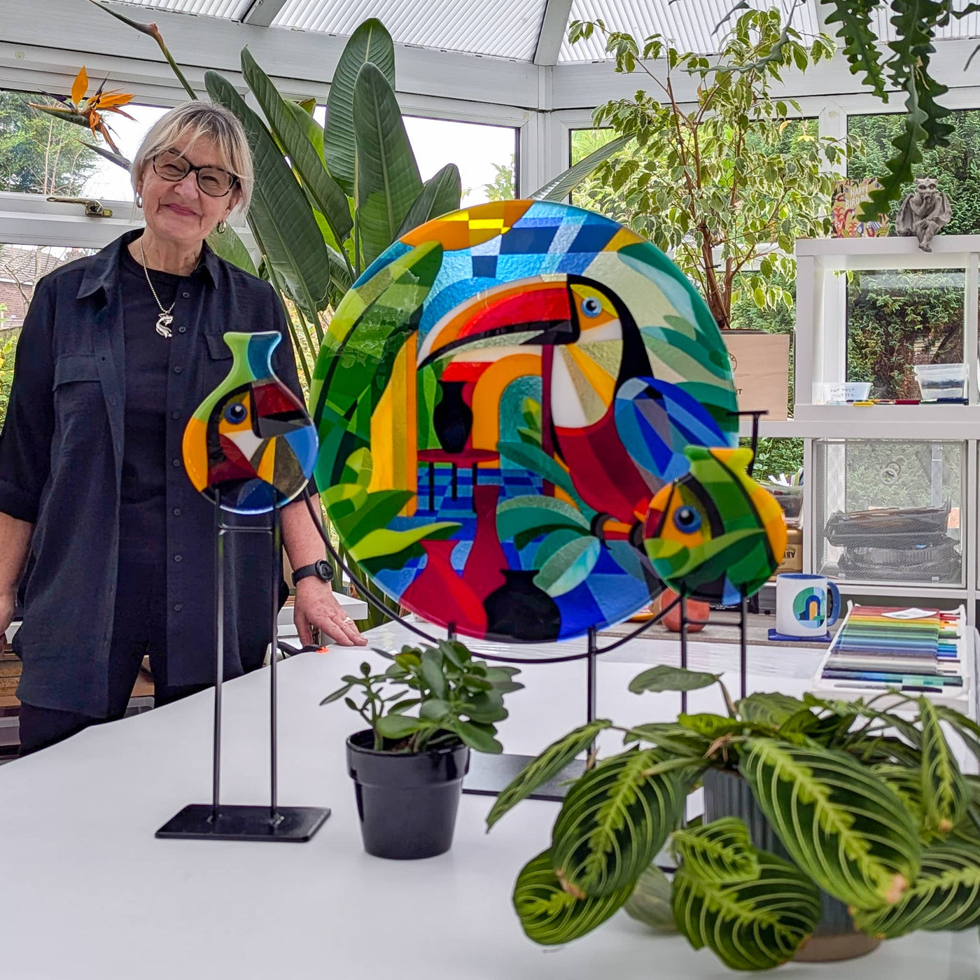 Linda Rossiter with the completed 'Toucans' contemporary Hard Edge glass roundel and two matching toucan miniatures displayed on their steel stands in her studio