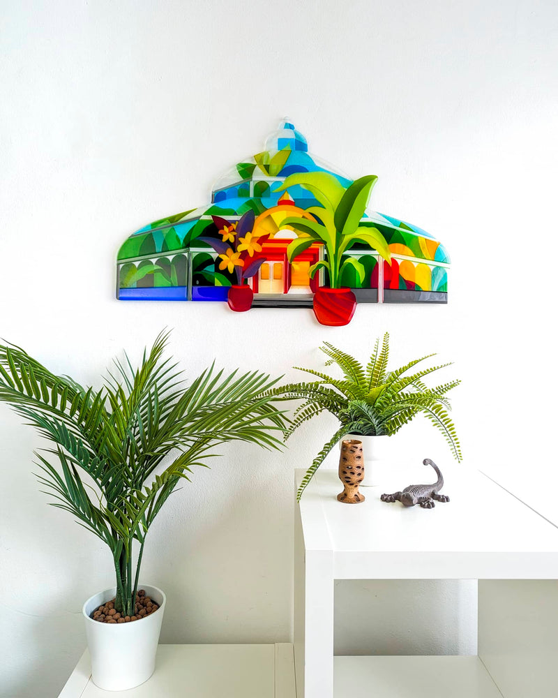 Palm House fused glass artwork displayed on a white wall above a table and indoor plants