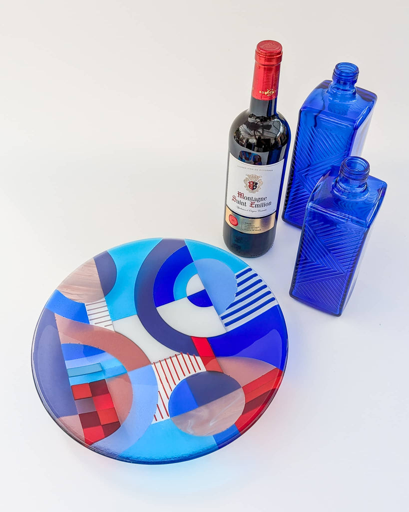 Fused glass bowl with bold Hard Edge geometric patterns in red, blue, and white, styled with a bottle of Montagne Saint-Émilion wine and two cobalt blue glass bottles on a white background