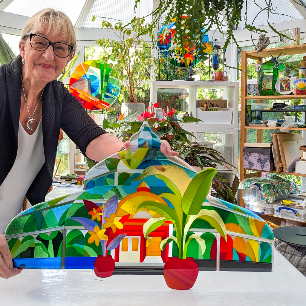 Linda Rossiter presenting 'The Palm House' fused glass artwork in her light-filled studio, with tropical plants and colourful glass designs in the background.