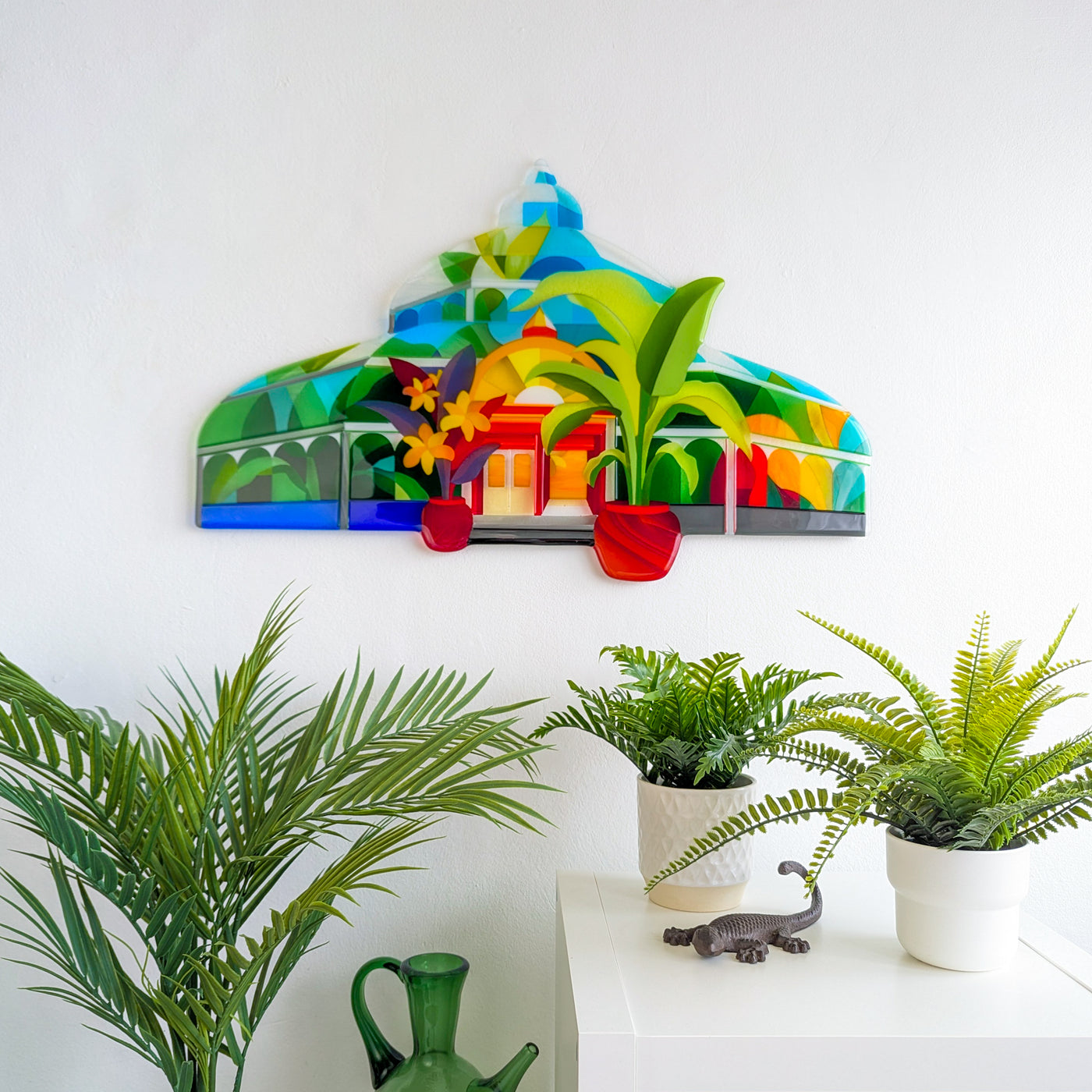 'The Palm House' fused glass artwork mounted on a white wall above a display of tropical plants and ceramics, styled in a bright modern interior.