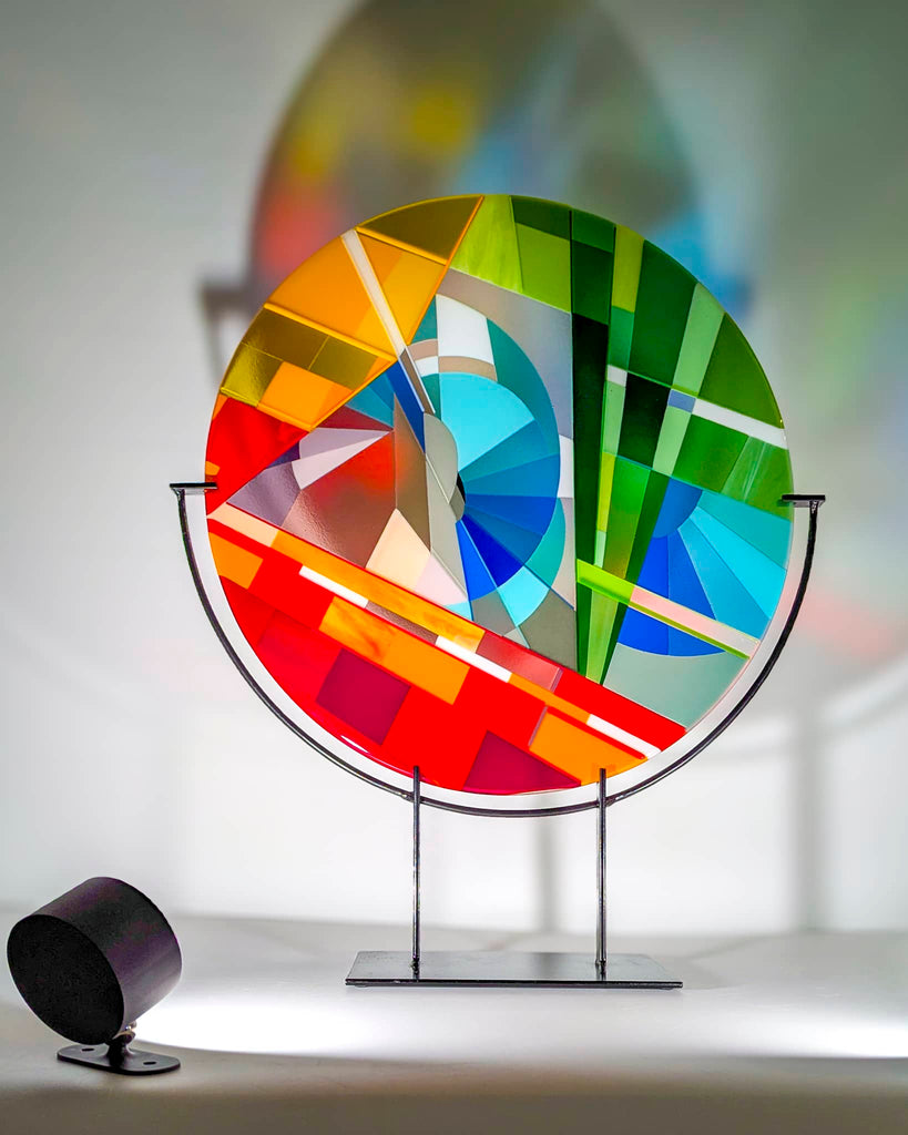 Colourful circular fused glass artwork on a stand illuminated by a small black USB spotlight – part of the premium gift package from Glass Art by Linda for commissions over £1,000