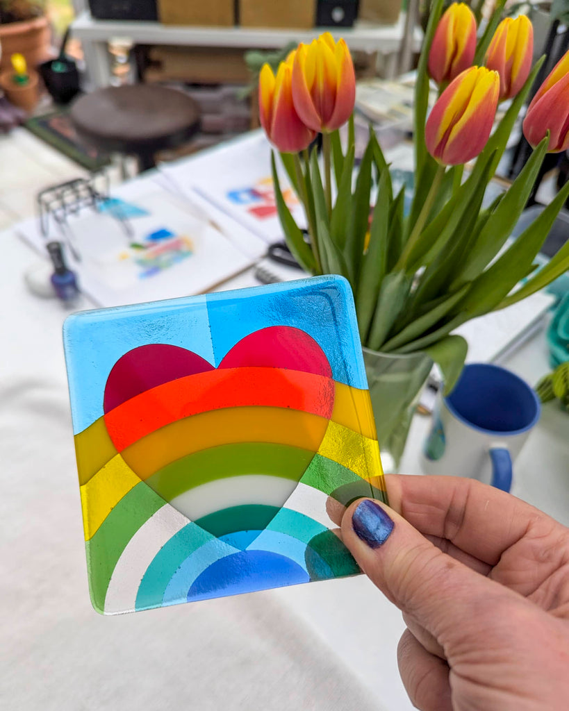 Fused glass miniature with a red heart above a layered rainbow design, held in hand in a bright glass art studio next to a vase of red and yellow tulips